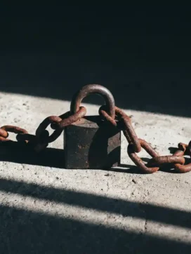 Roestig ijzeren hangslot omwikkeld met gebroken kettingen op betonnen vloer, dramatisch zijlicht werpt scherpe schaduwen.