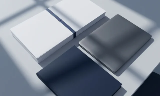 Stapel witte compliance-documenten naast een laptop op een lichtgrijs bureau, met een donkerblauwe map als ankerpunt.