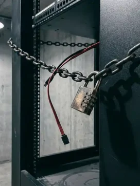 Stalen ketting met cijferslot om een serverrack, doorgesneden kabel bungelend, tegen betonnen muur in dramatisch tegenlicht.
